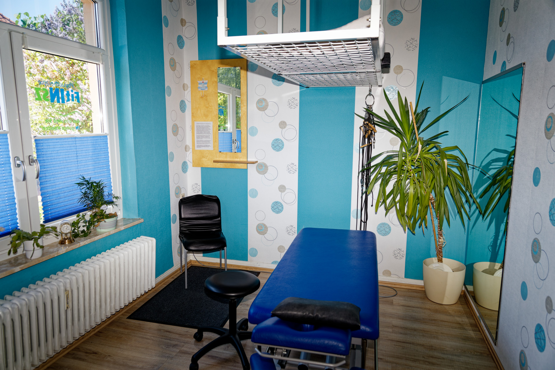Behandlungsraum eins mit einer Behandlungsliege und dadrüber mit dem Gitter vm Schlingentisch, Ein Stuhl zum umziehen ist daneben sowie eine Pflanze